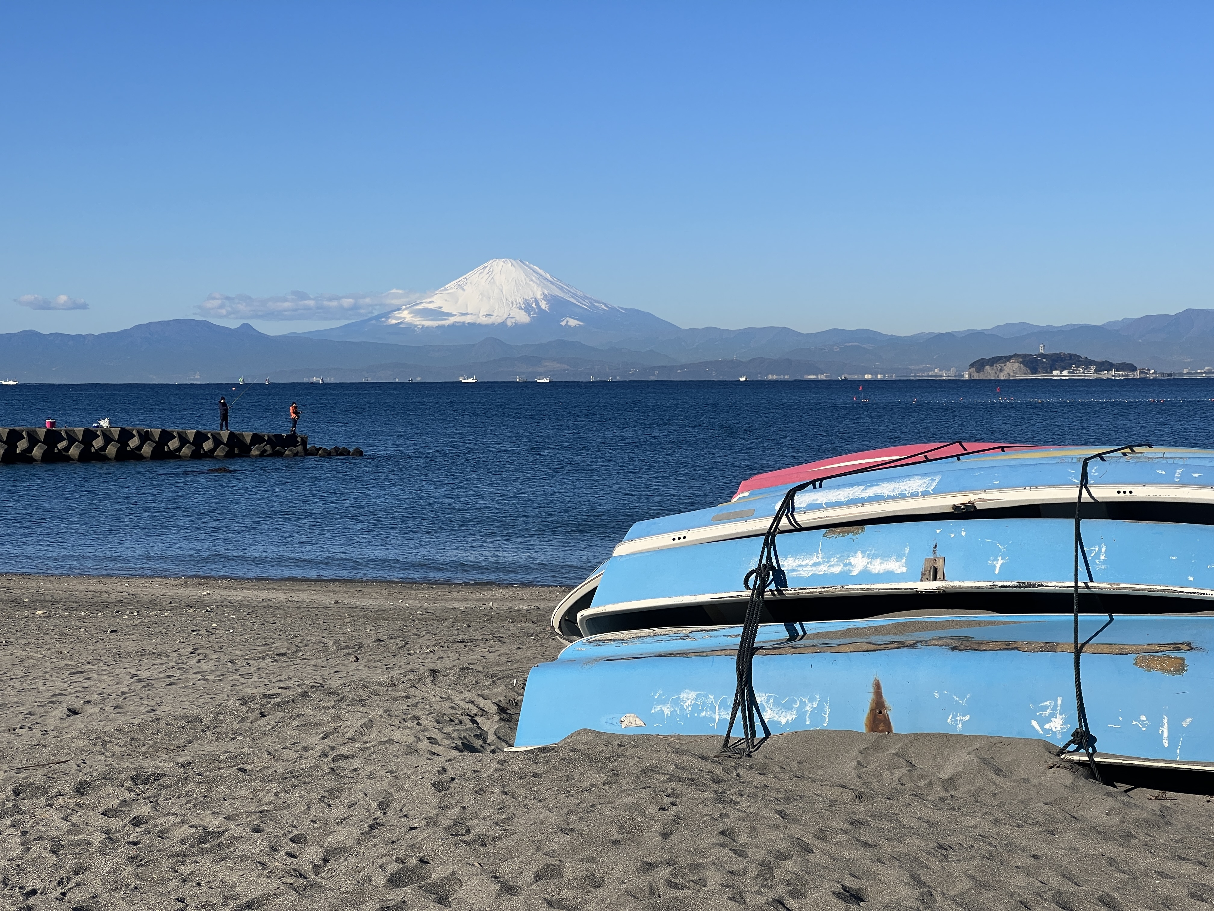 A peaceful beach scene with Mount Fuji in the distance, row boats in the foreground and people fishing from a break wall